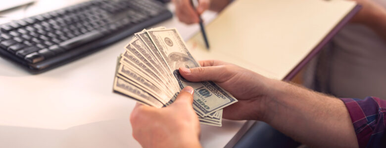 A person counting a stack of U.S. dollar bills at a desk, symbolizing financial transactions or cash handling. In the background, a woman is writing on a clipboard, and a keyboard and office setup are visible, indicating a work or business environment.